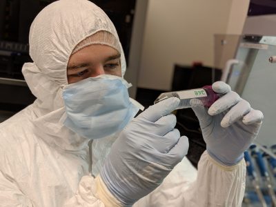 A man wearing protecting personal equipment, including a mask over the loer part of his face, looking at a small tube containing a sample of sediment 