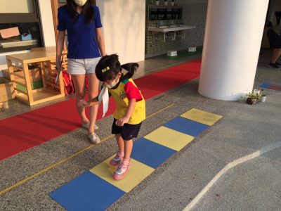 A child jumps on flat blue and yellow foam squares on the ground. She is getting her coordination assessed by a researcher who stands beside her