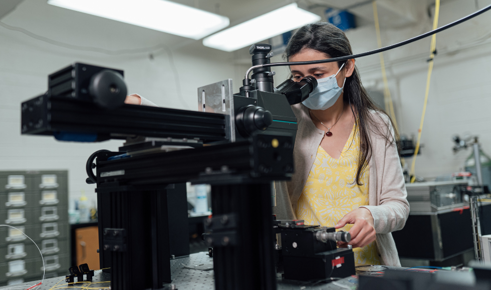 A photo of Engineering Physics student PhD student Khadijeh Miarabbas Kianii. She is wearing a mask and looking into an ocular lens that is attached to a large piece of machinery that is sitting on top of a table.