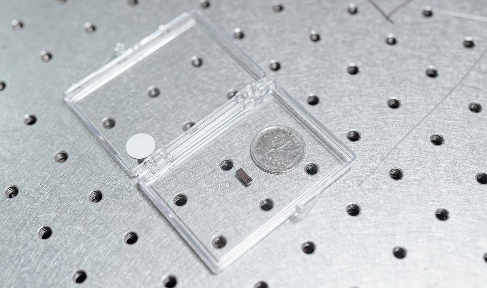 A photo of a plastic case on a tabletop containing a small, silicon chip with a Canadian quarter beside it to demonstrate size. The chip is about an eighth the size of the coin. 