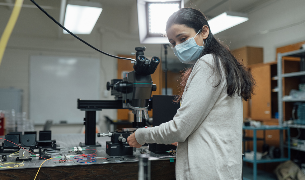 A photo of Engineering Physics student PhD student Khadijeh Miarabbas Kianii inside a labratory. She is wearing a mask and looking back over her shoulder. In front of her there is a large piece of equipment with several ocular lenses. 
