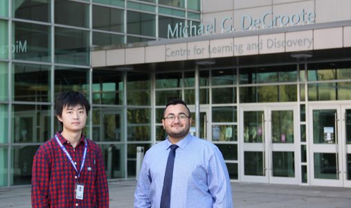 McMaster students Hugo Yan and Kian Yousefi Kousha standing in front of the Michael G. DeGroote Centre for Learning and Discovery on McMaster's campus. Hugo Yan is wearing a red and blue checkered shirt with a lanyard hanging from his neck. Kian Yousefi Kousha is wearing a light blue dress shirt and a dark blue tie. 
