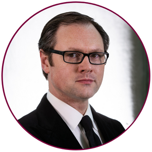 A headshot of James MacKillop. He is wearing glasses, a white dress shirt, black tie and black suit jacket. He is looking directly at the camera without smiling. 