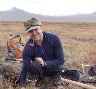 A smiling man with a pipe in his mouth crouching down with a backpack outdoors.