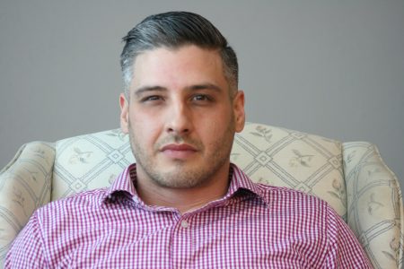 A man sits looking at the camera weaing a red gingham shirt