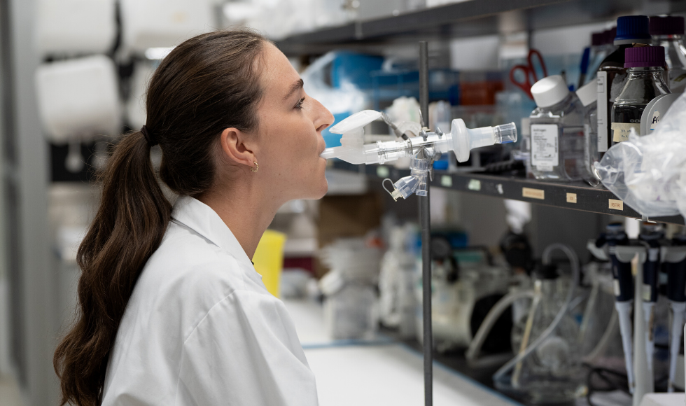 A woman in a laboratory setting wearing a white lab coat, inhaling through a clear plastic tube in her mouth, like a straw.