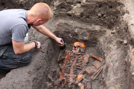 A researcher holding a brush in one arm, leaning over into a shallow pit that contains a skeleton 