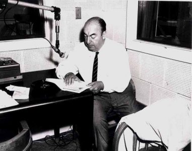 A black-and-white photo of Pablo Neruda sitting at a desk, looking down at a book in front of him. There is a microphone hanging from a suspended bar in front of him. 
