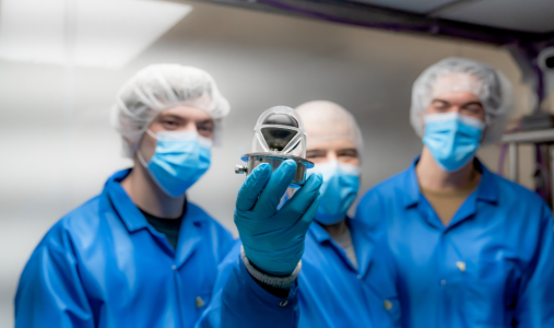 3 scientists in full lab protective gera and masks. The one in the centre is holding up a small device to the camera. That's the radiation detecting part of NEUDOSE