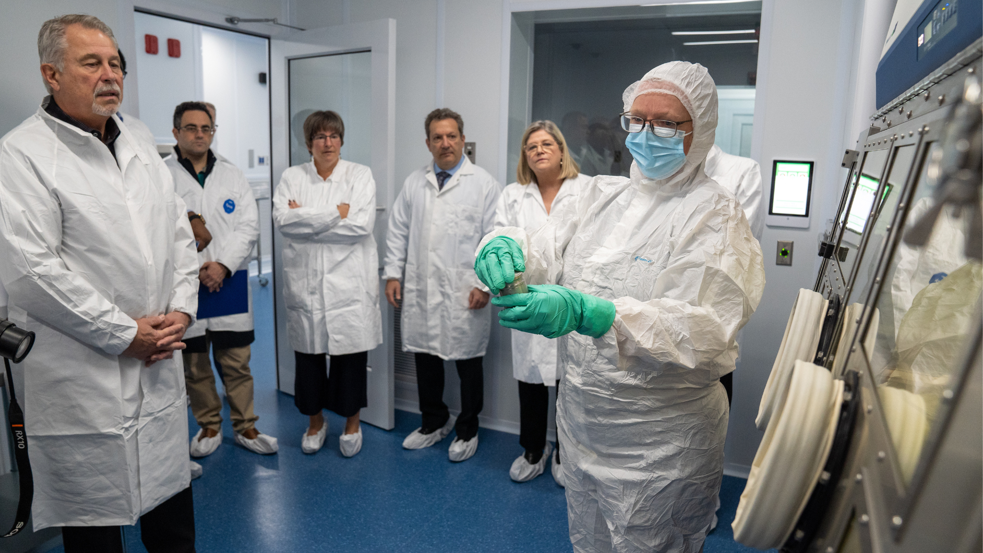  Five people wearing lab coats look on as someone in full personal protective equipment makes a demonstration in front of laboratory equipment 