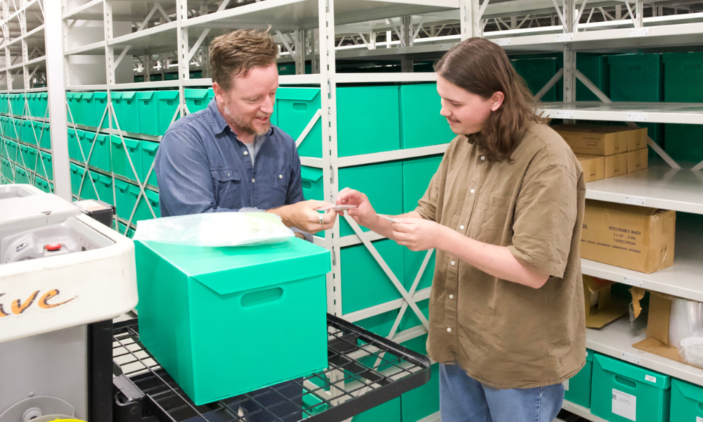 McMaster professor Andrew Roddick and student Rylan Godbout examining a slide. They are in a warehouse setting with numerous green boxes around them. 