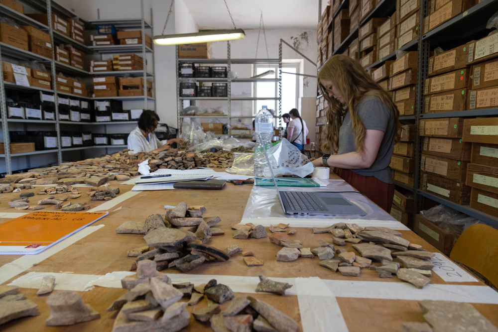 A photo of McMaster student Christine Davidson as she sorts and digitally catalogs a number of pottery sherds laid out on a large table in front of her.