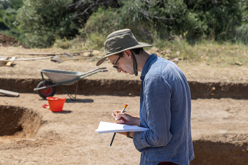 A photo of PhD student Eric Del Fabbro on site at the Metaponto Archaeological site. He is taking notes and standing in a hole in the ground.