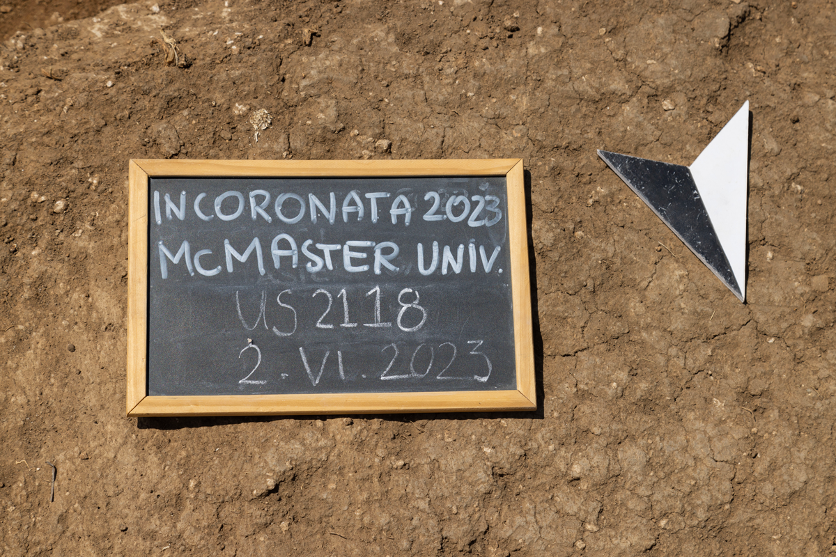 A sign lies on the ground of the Metaponto Archaeological Site. It reads "Incoronata 2023 McMaster Univ. US2118 2.VI.2023"