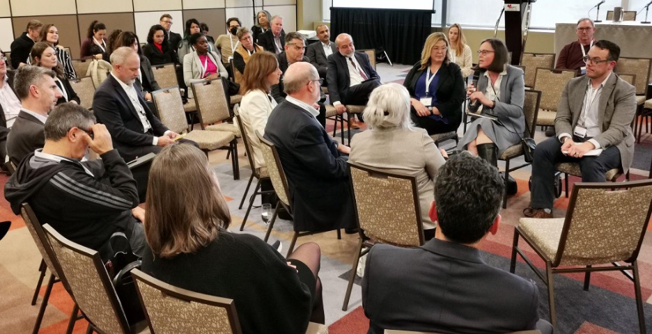 A large number of people sitting in a circle and on the perimeter of the circle at a "fishbowl" panel discussion.