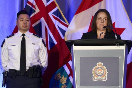 Image of an officer standing beside a podium, while a person talks on the microphone 