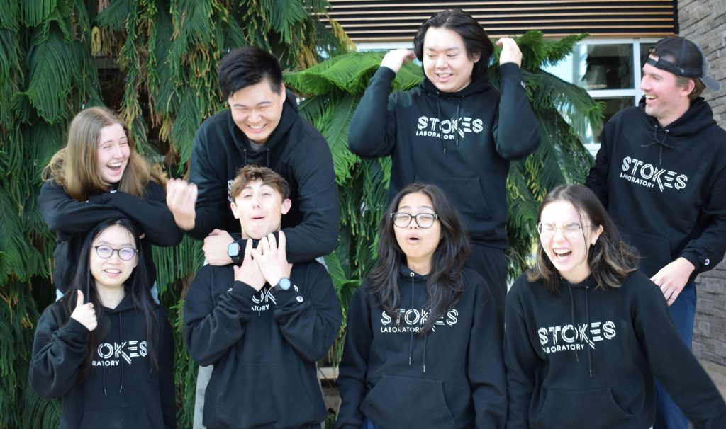 Eight students in "Stoked Bio" sweatshirts laugh and clown around in a group photo