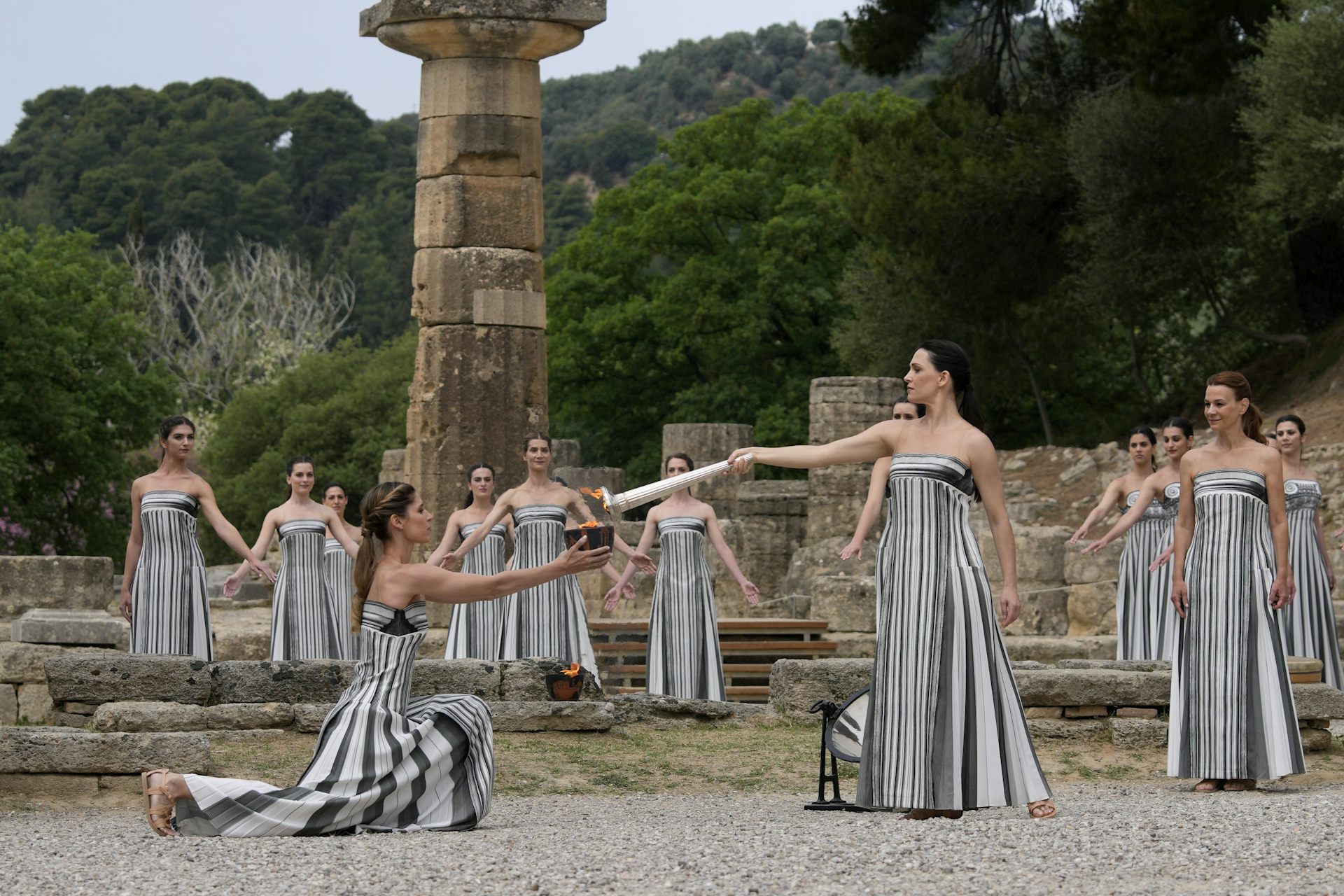 "A group of women wearing dresses stylized to look like Greek columns stand among ancient ruins. One of the women kneels and holds a bowl in her outstretched arms while another lights the bowl with a torch"