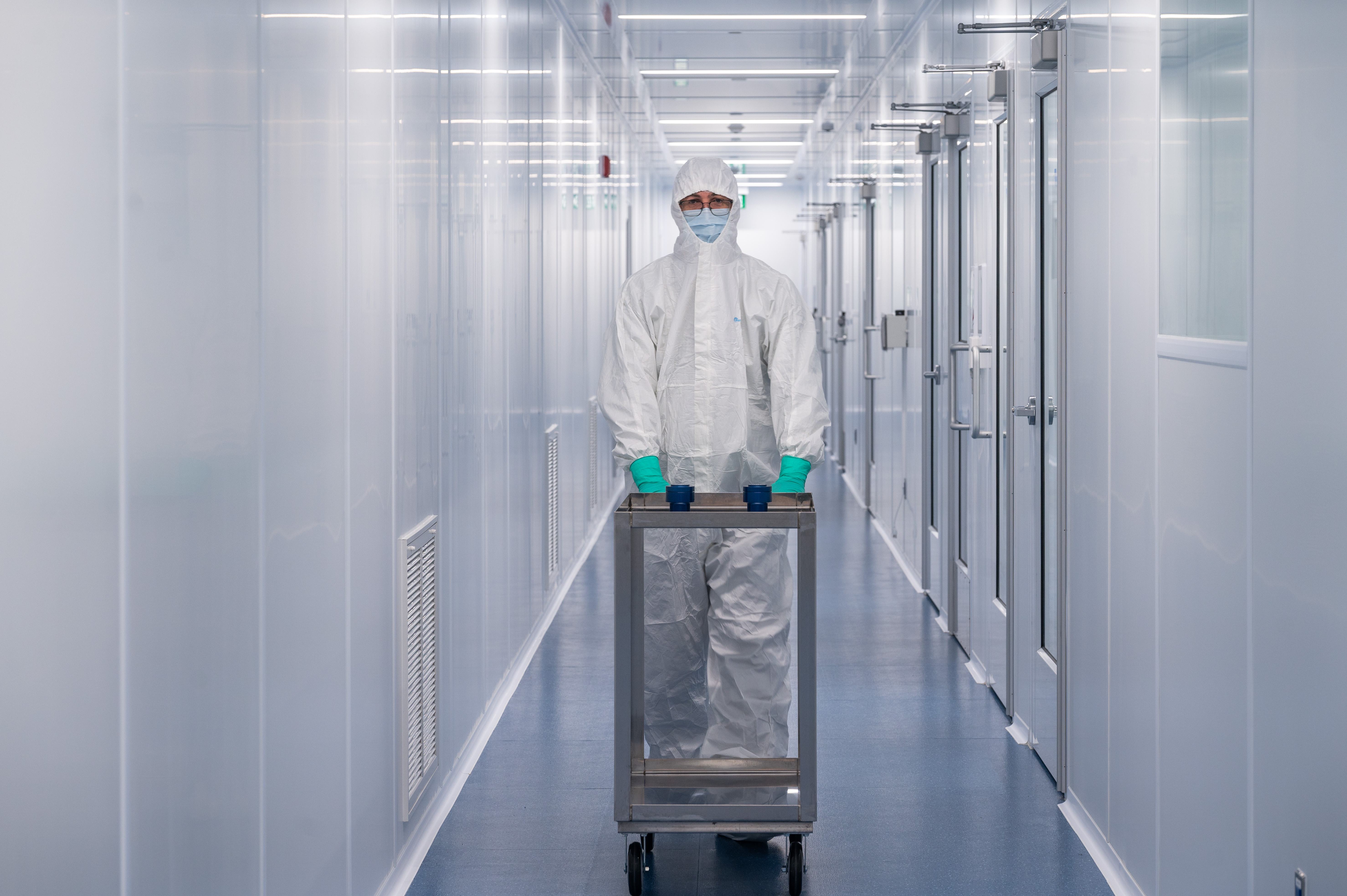 A person in full protective lab coveralls, a mask and gloves, wheels a cart with Fusion Pharmaceuticals vials on it down a white hallway.