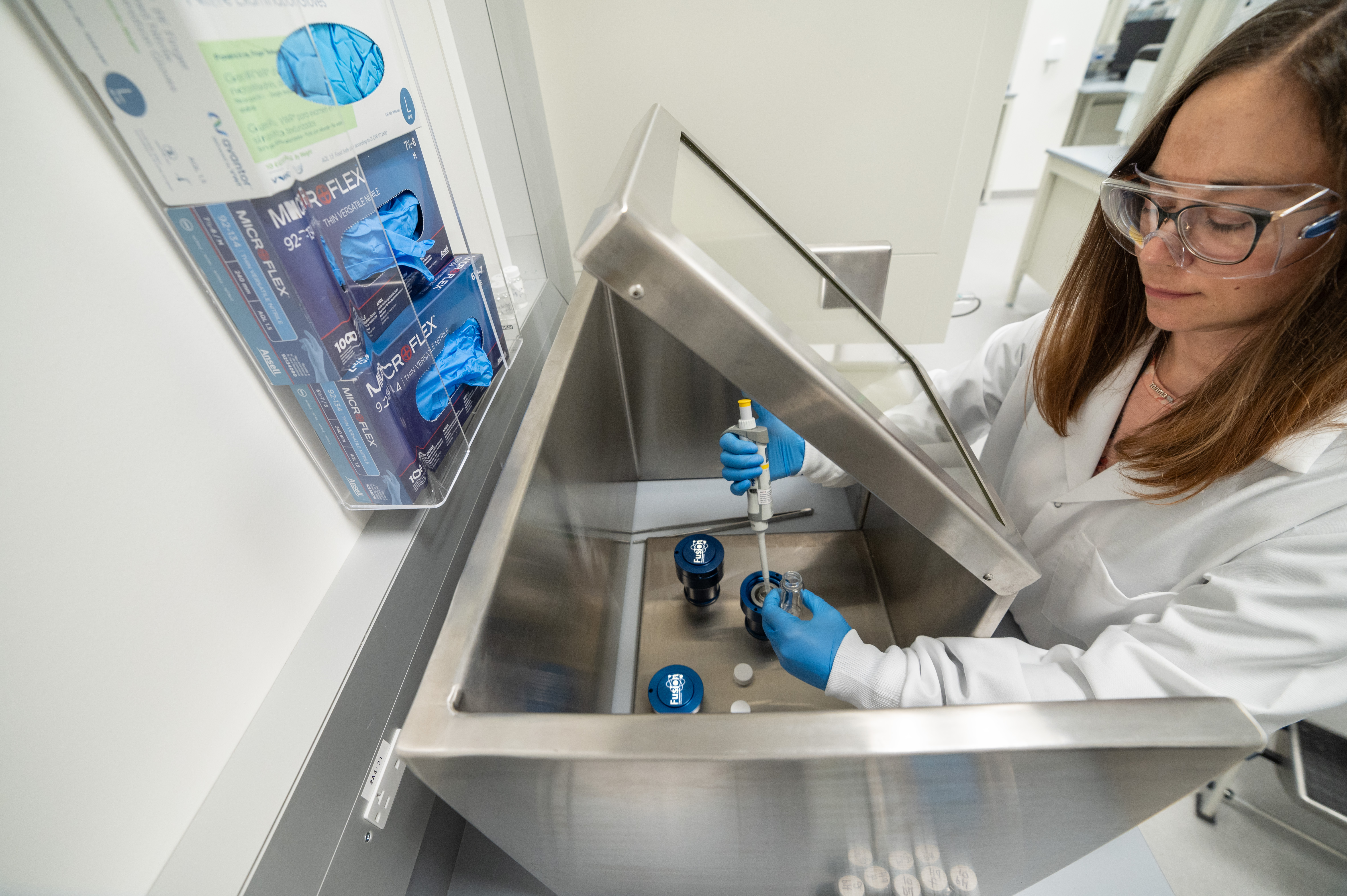 Seen from above: A person with long dark hair in a lab coat and gloves is working with vials and bottles labelled Fusion Pharmaceuticals in a lab setting.