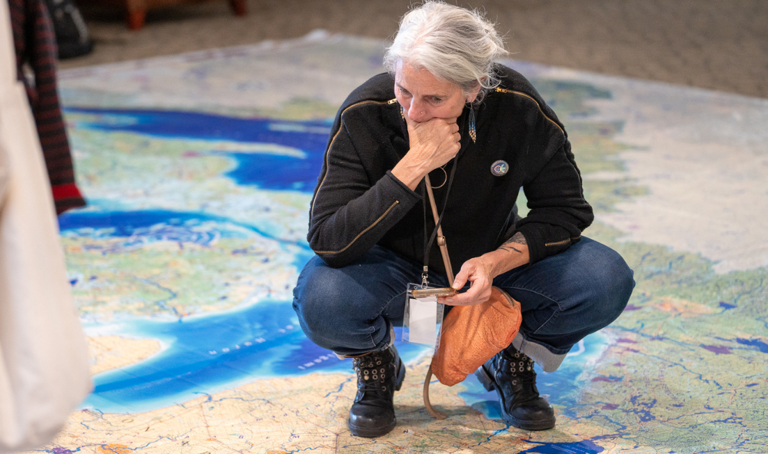 A person with grey hair, a black sweater and jeans crouches down holding their phone above an enormous floor map showing the Great Lakes watershed.