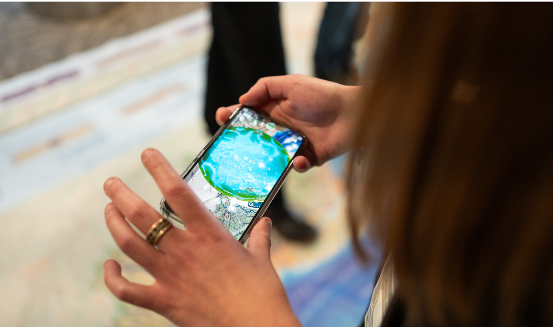 A closeup of a smartphone opening augmented reality features from the giant floor map of the Great Lakes watershed.