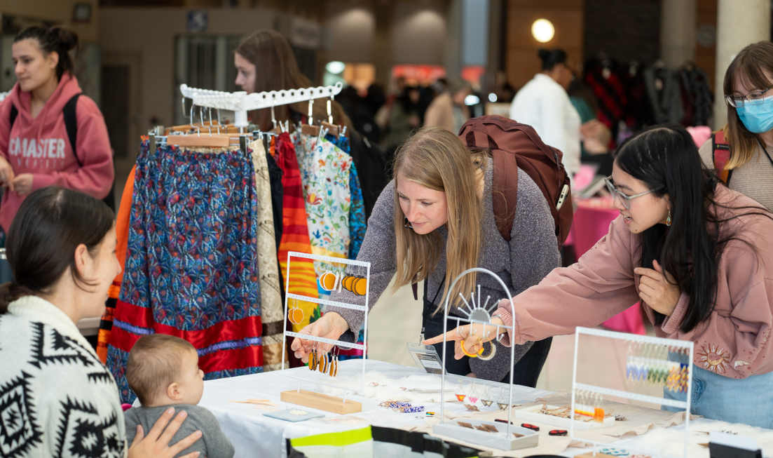 A vendor holding a baby smiles at two people who are looking at the earrings for sale at their table at the Indigenous Marketplace in MUSC.