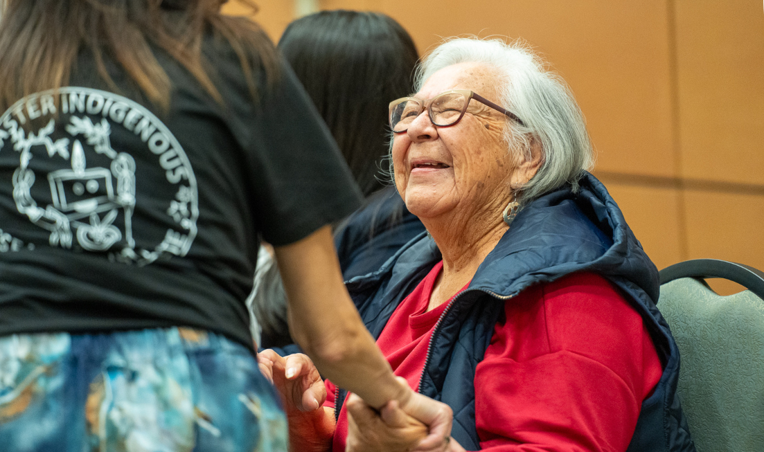 Faithkeeper and Elder Norma Jacobs smiles and holds the hand of someone who has their back to us.