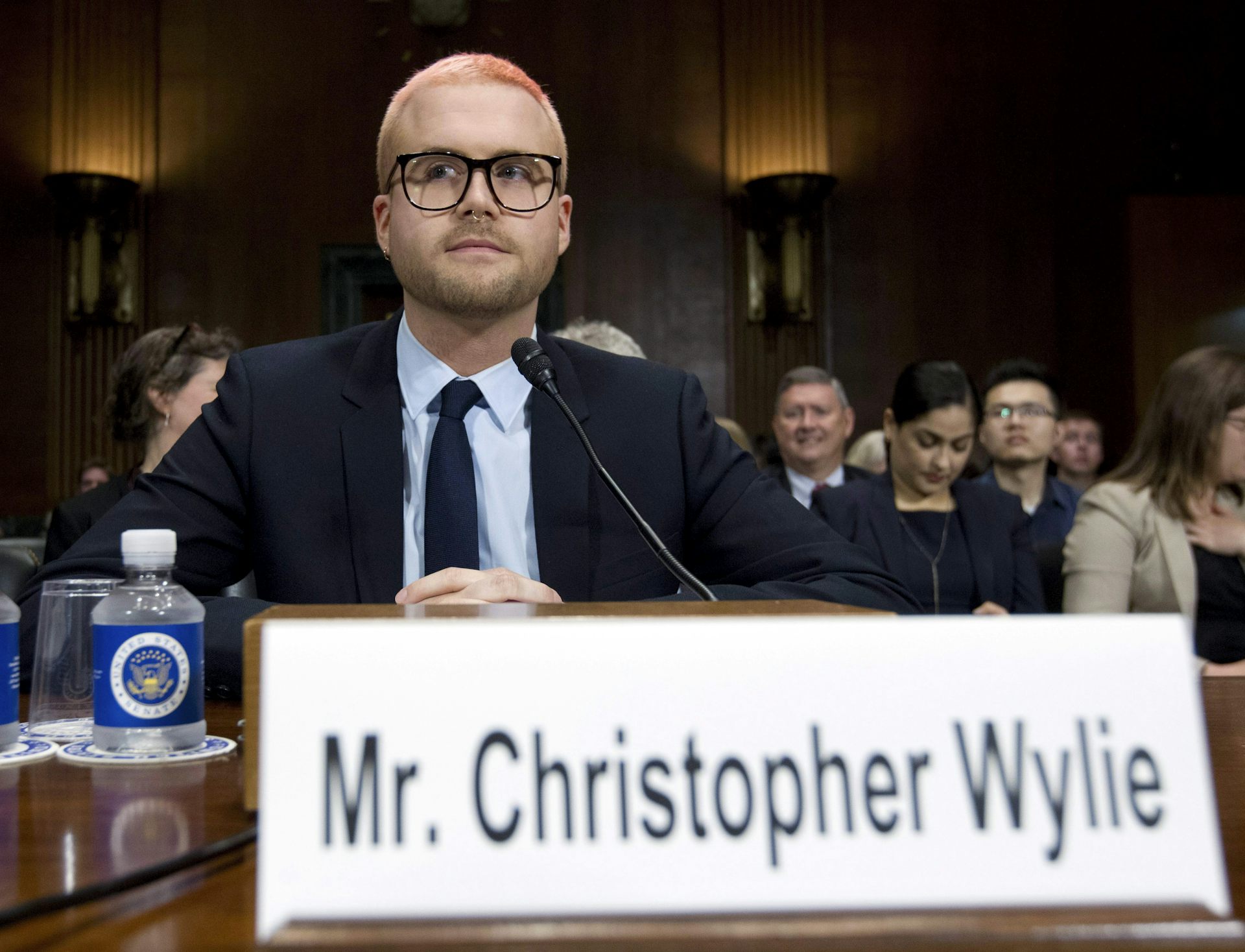 A man with a buzzcut and glasses testifies at a hearing. His name, Christopher Wylie, is on a name plate in front of him.