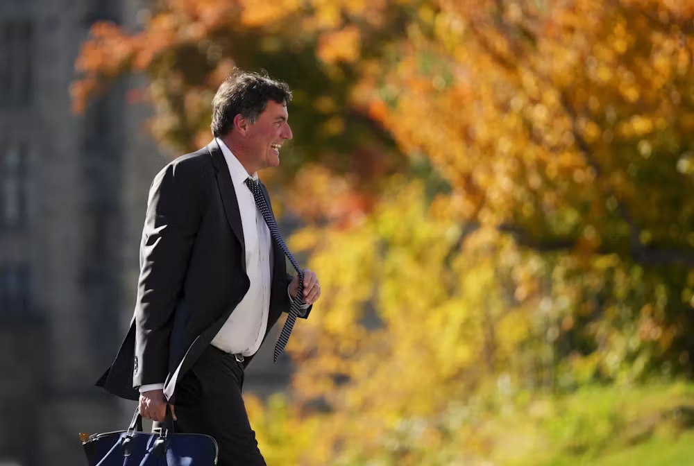 A man with brown hair walks outdoors as trees in full fall colour appear beside him.