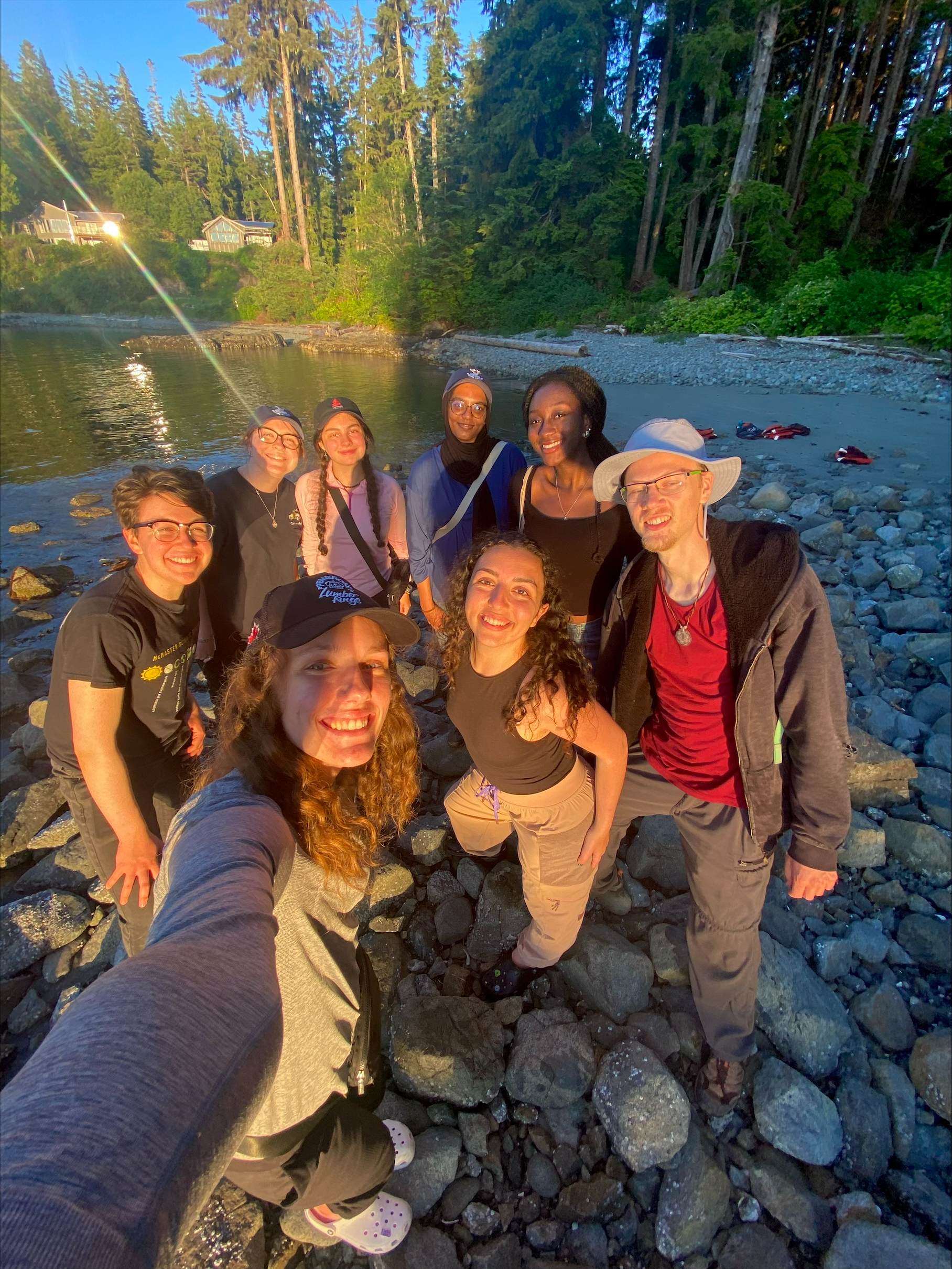 Eight smiling students look upward to take selfie in golden yellow sunset on a rocky beach.