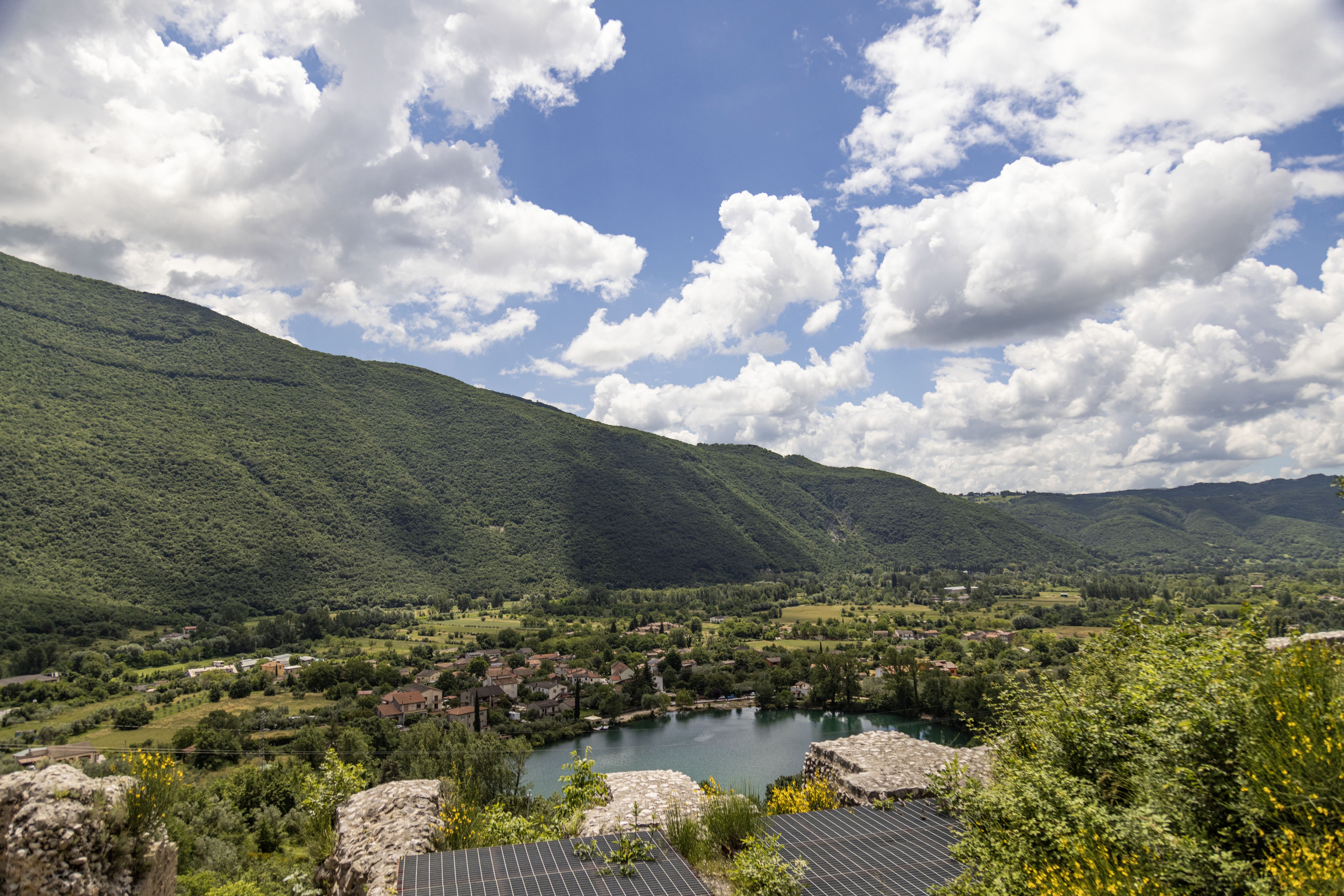 A stunning view of a lake in a valley, surrounded by green hills, under a blue sky with fluffy white clouds on a sunny day.