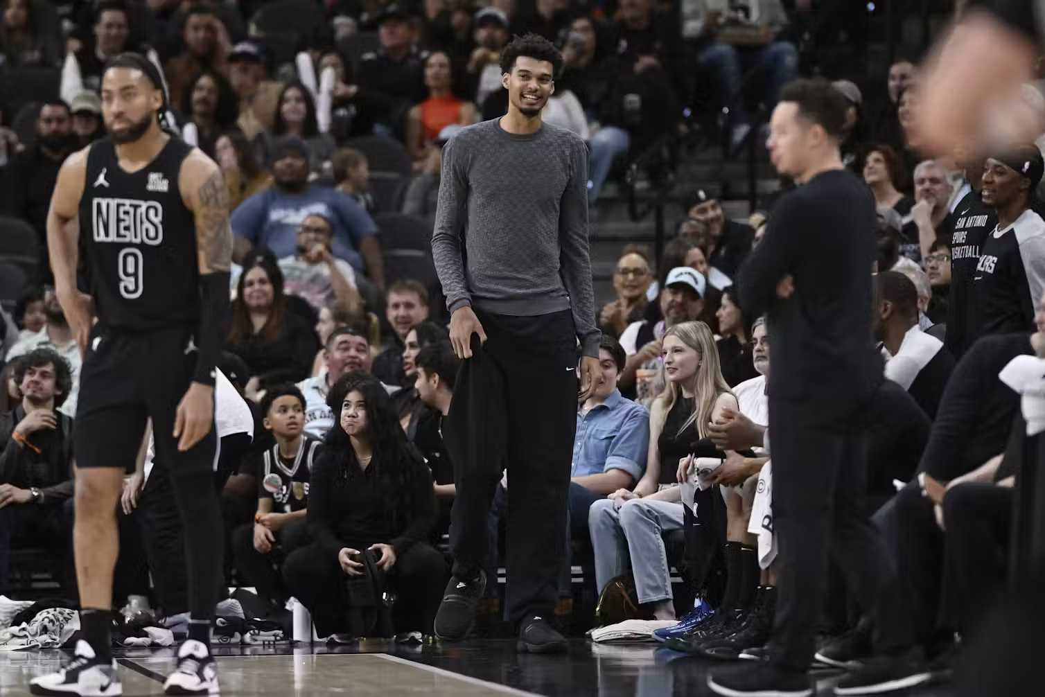 The sidelines of a basketball game, with several people sitting and a few people standing up.