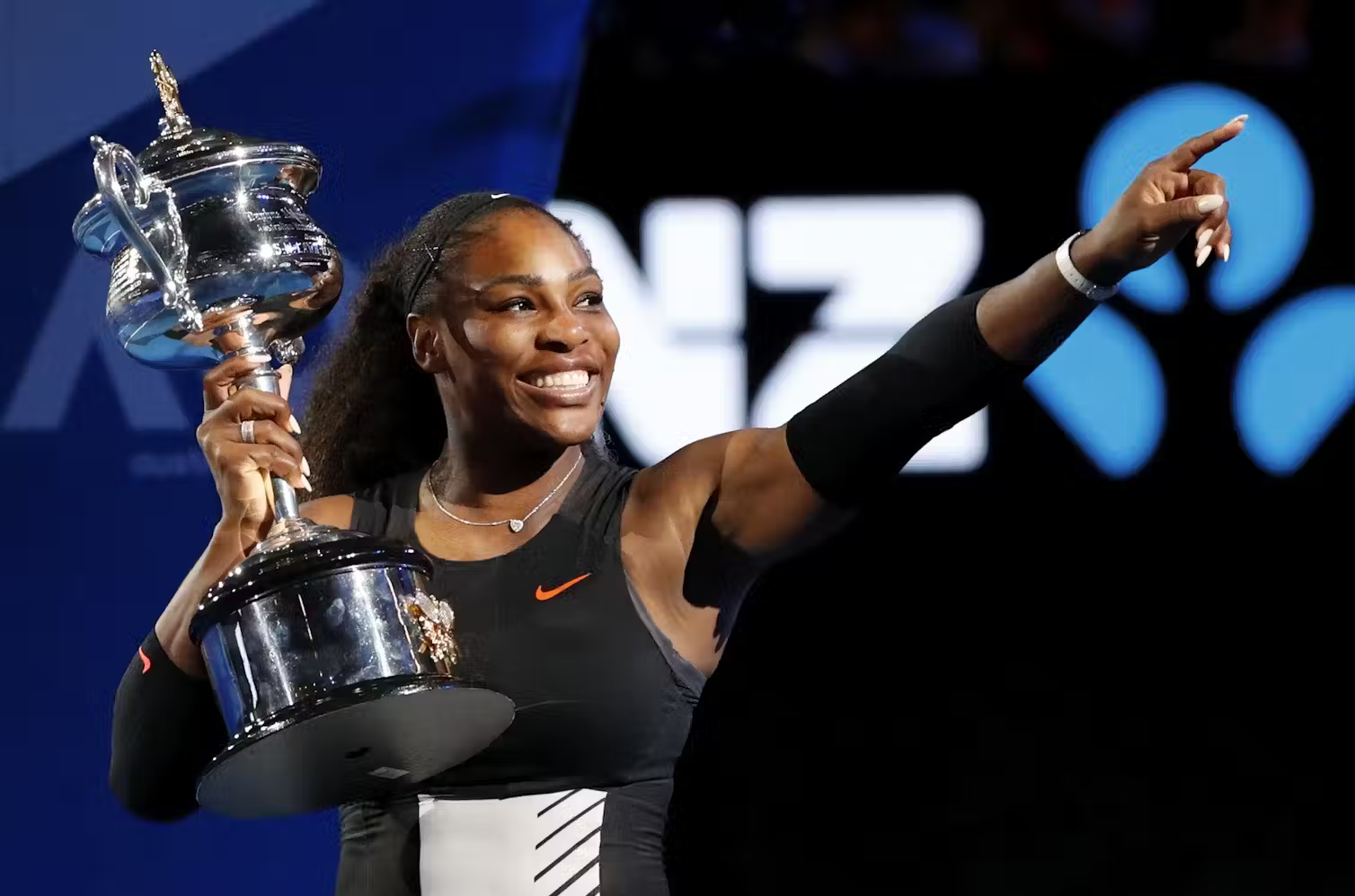 Serena Williams holds a trophy and smiles, pointing up and to her left.