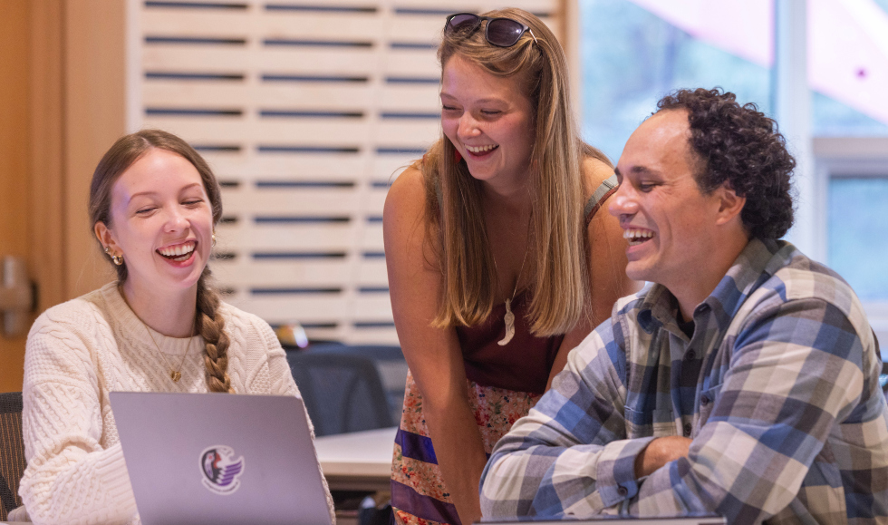 Two people sit at a table, and one person, standing, leans over. They are all looking at a laptop that sits on the table, and they are laughing.