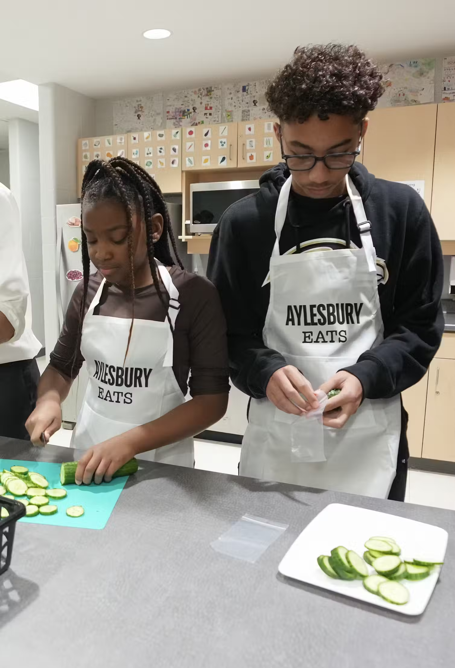 Two teenaged children wearing aprons prepare cucumbers in a kitchen.