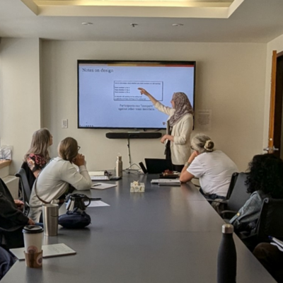 Several people sit around a large meeting table, all looking at a screen on the wall, with one person pointing to something on the screen.