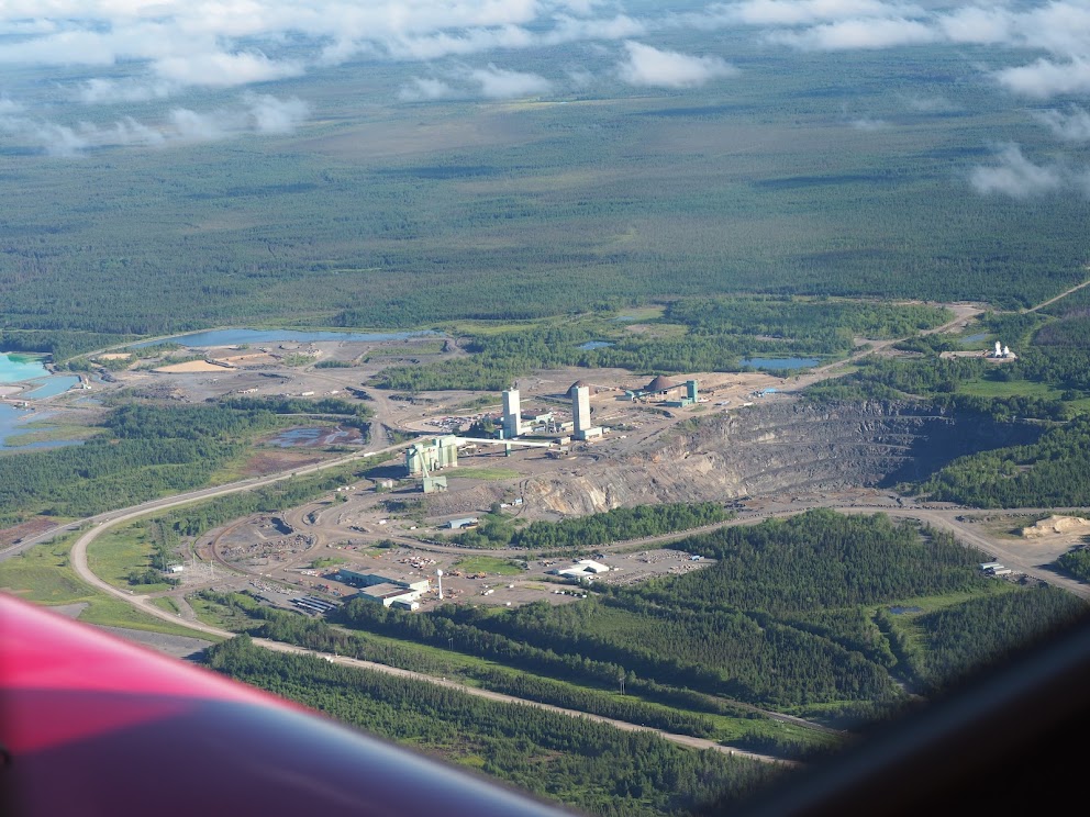 An aerial shot from a plane, of a mining operation below. A large mining pit is visible, surrounded by greenery all around.