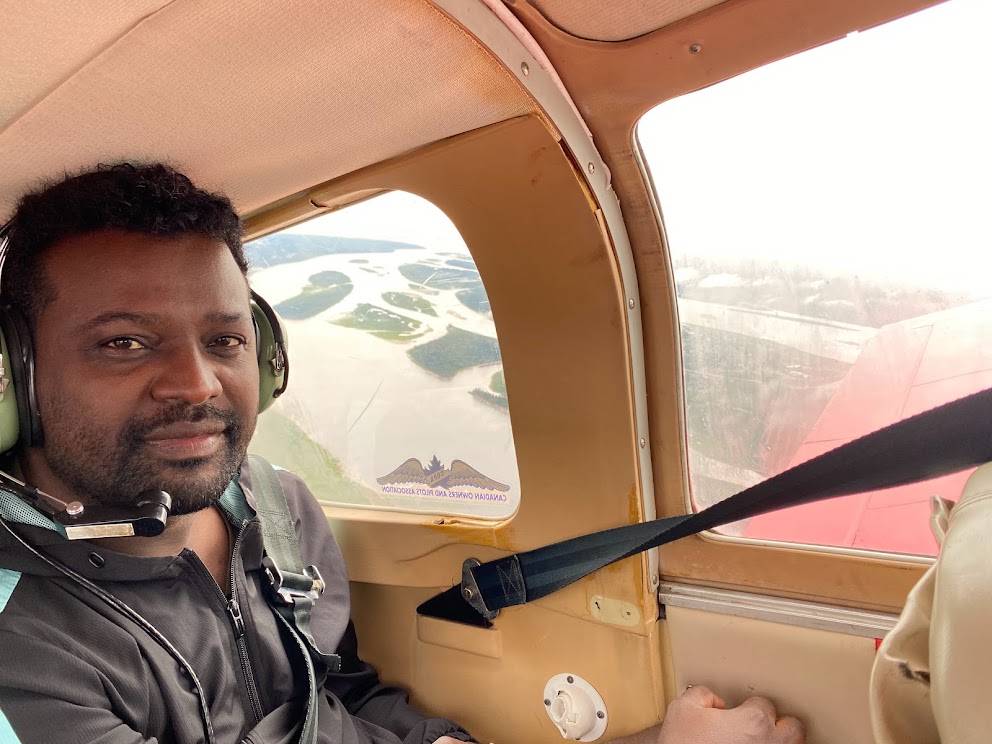 A man sits in a small airplane. The airplane window shows greenery and water.