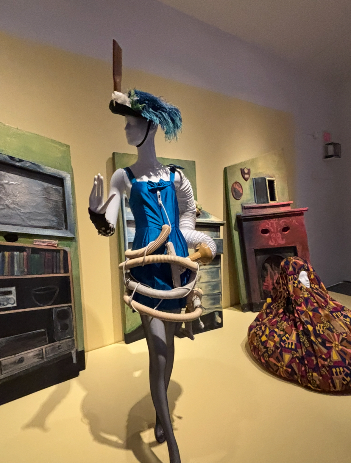 A mannequin stands in an artistic rendering of a sitting room, dressed in a feathered hat, blue dress and tubing materials.