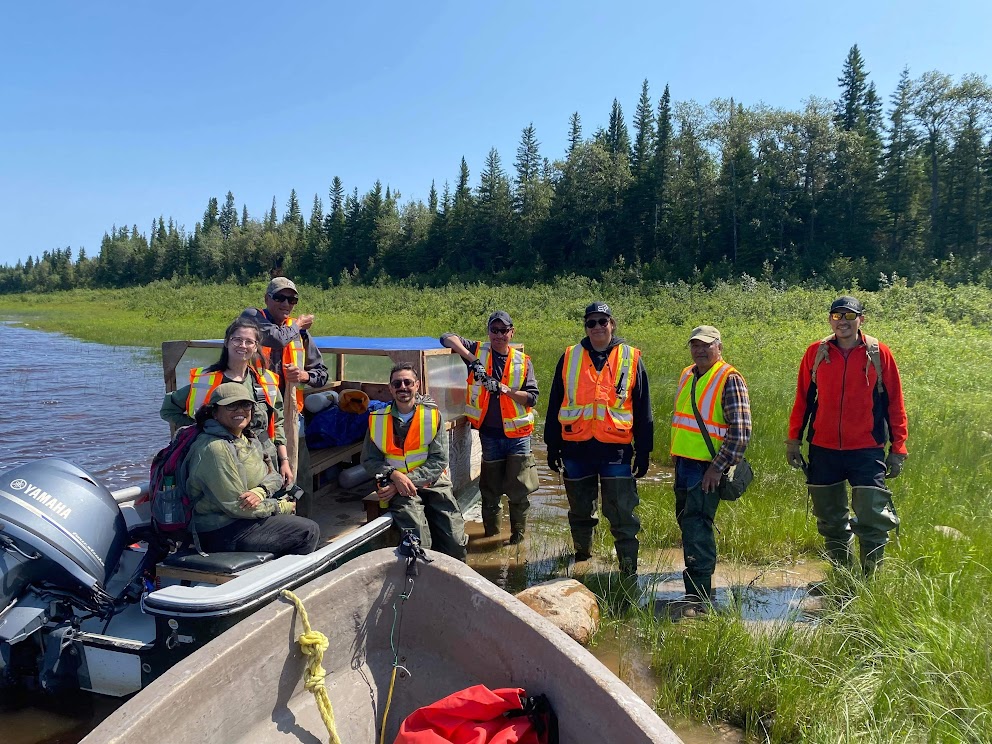 A group of seven people stand beside a boat, which is sitting in the water on the edge of a green marsh. All of the people are wearing orange safety vests.