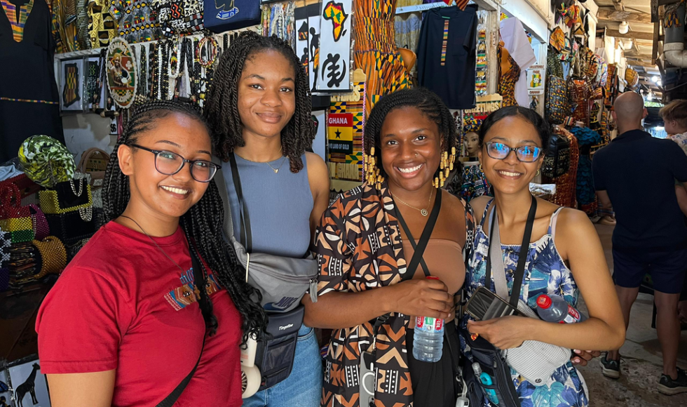 Four women, dressed casually, stand together, with a bright and colour background behind them.