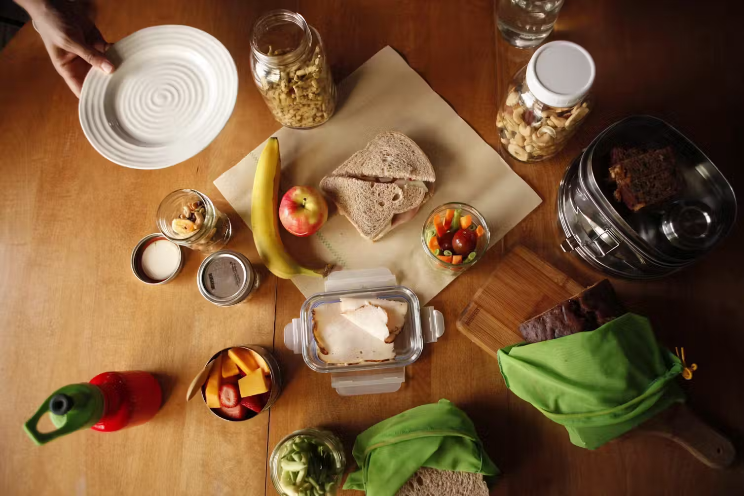 An overhead shot of a table, with reusable containers, wrappings, and food items including a banana, apple, and sandwich.