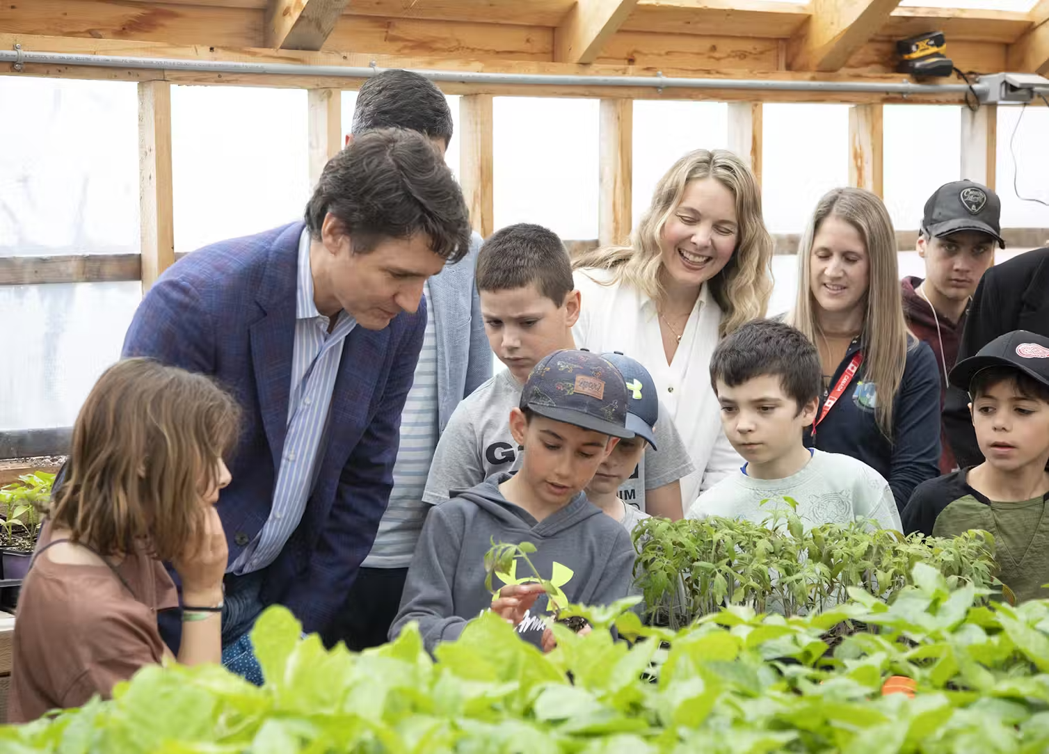 Former Prime Minister Justin Trudeau stands with kids in a greenhouse, looking at leafy plants.