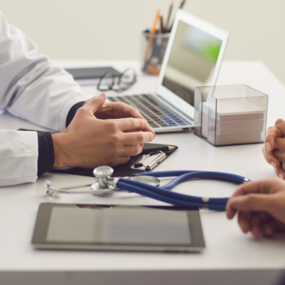 A desk with a laptop and stethoscope, with two people's hands resting on the desk from opposite sides of the side.