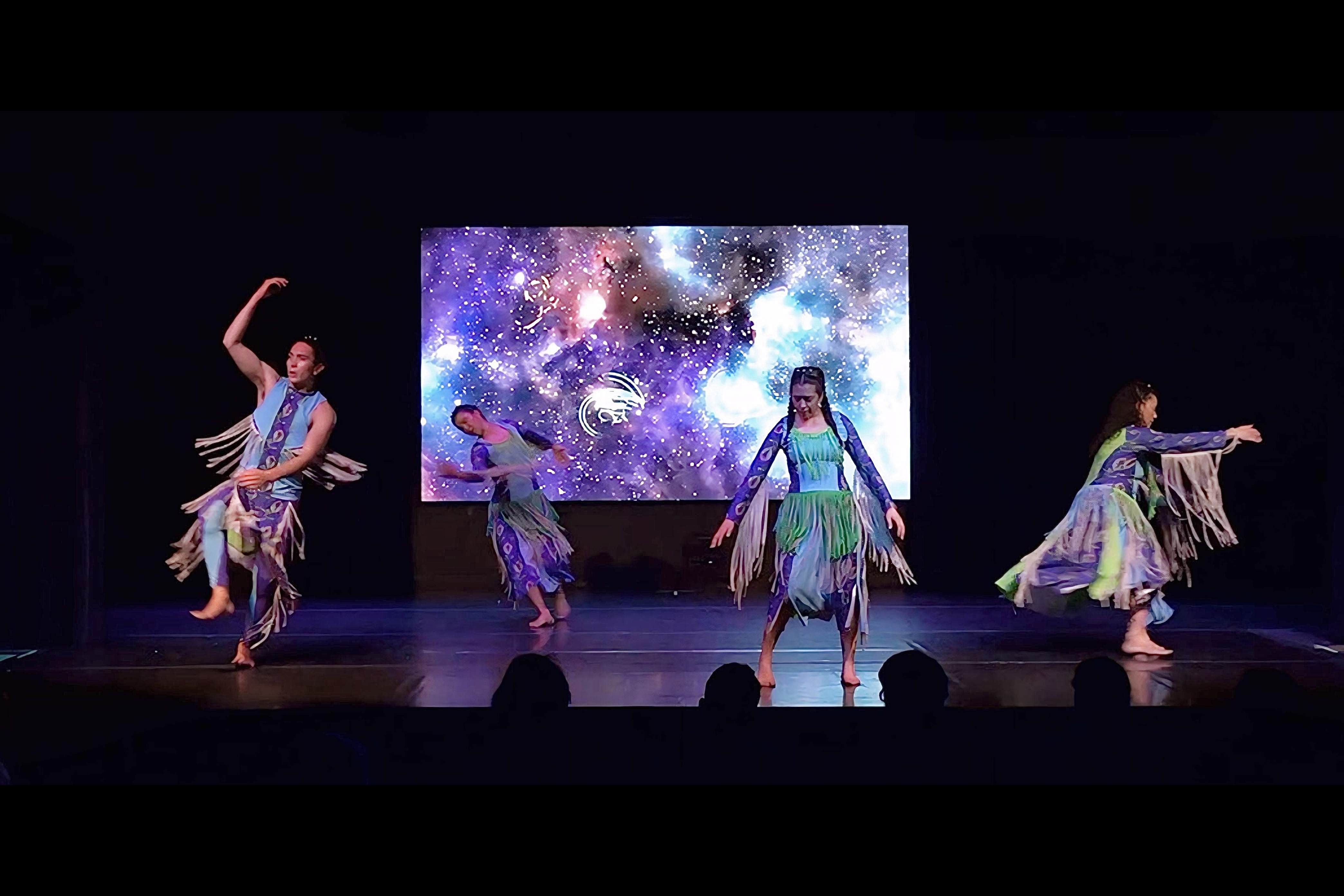 Four people dance on a stage, with a screen behind them showing stars and space.