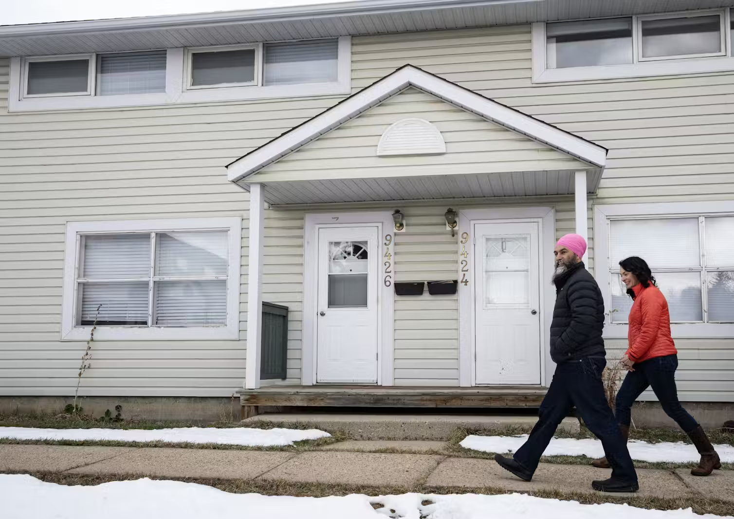 Two people, one a man wearing a bright pink turban, walk along a snowy residential street.