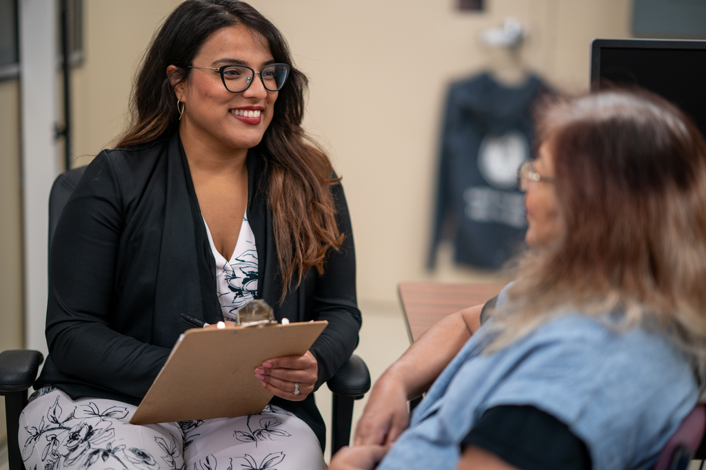 A woman sits with a clipboard, speaking to another woman.