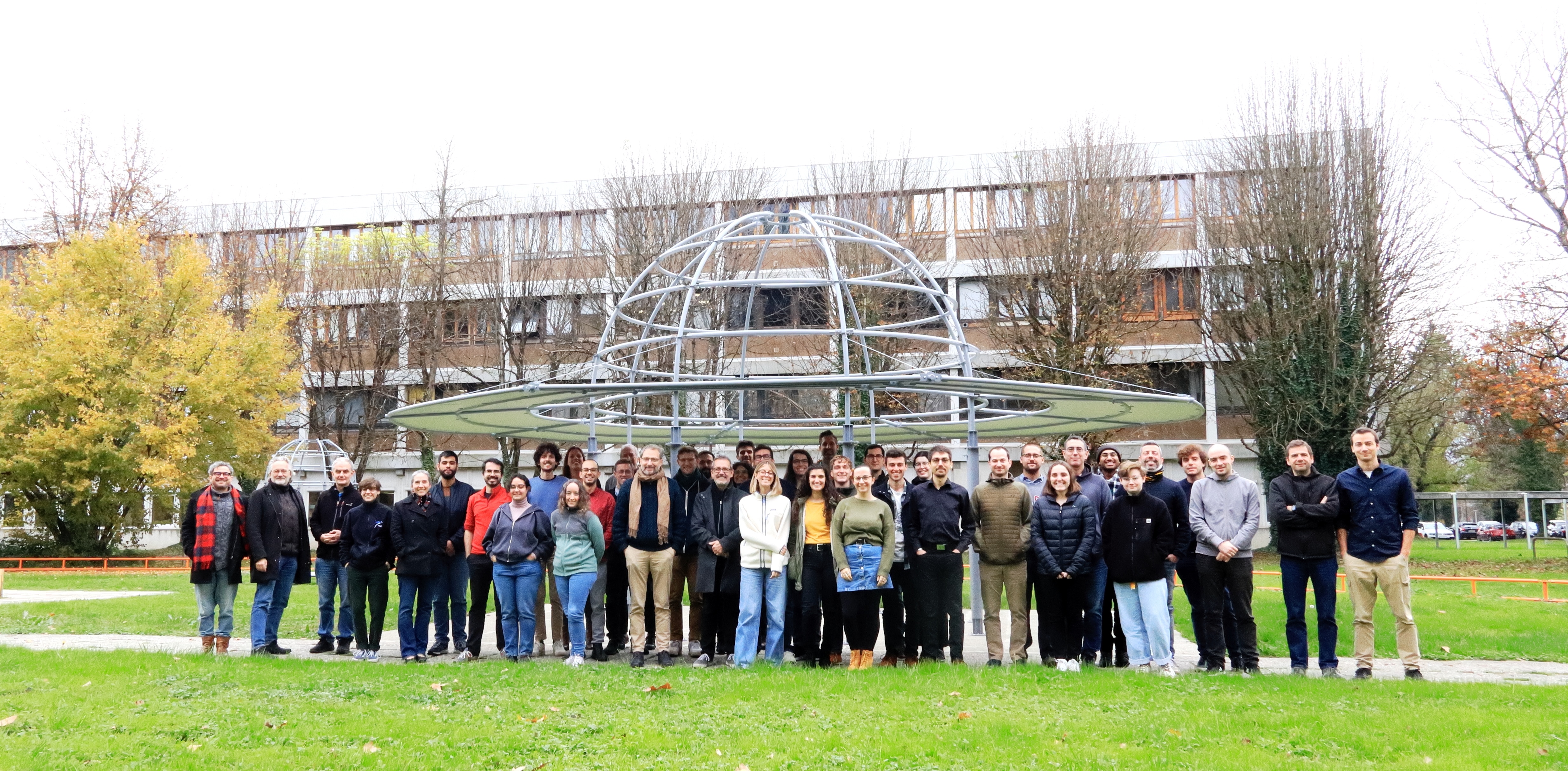 Wide shot of a group of a few dozen people standing side-by-side outdoors in front of an observatory building in jackets and sweatshirts.