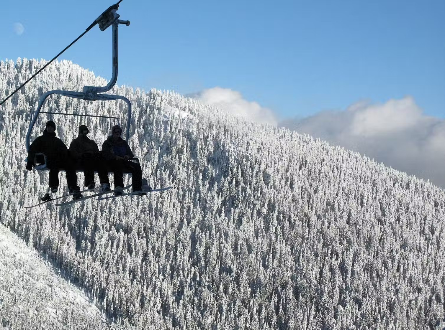 Three people sit on a ski lift, above snowy, tree-covered hills.